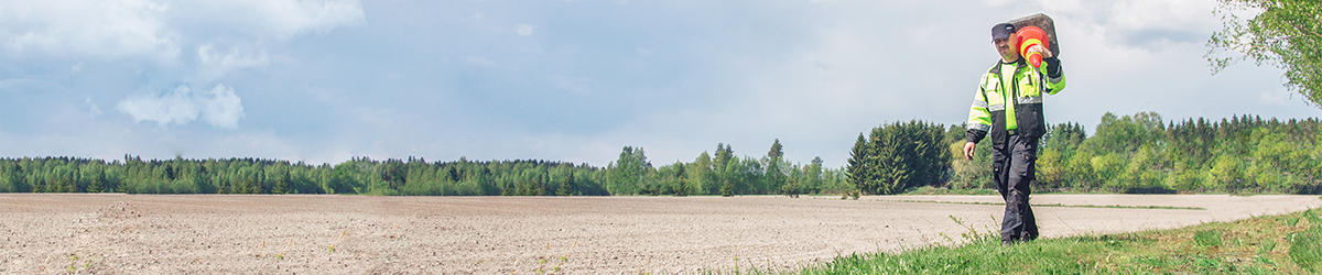 Huomiovärisiin vaatteisiin pukeutunut mies kantaa merkkitötteröä pellon reunalla.