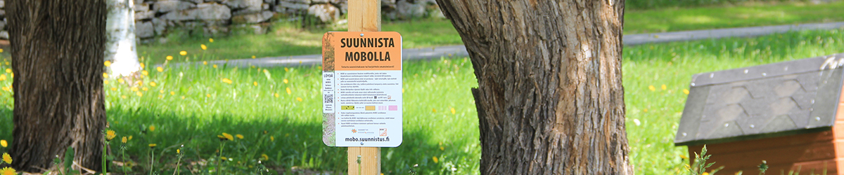 Mobo-suunnistuksen lähtöpisteellä oleva opaskyltti ulkona nurmikolla, puiden välissä.